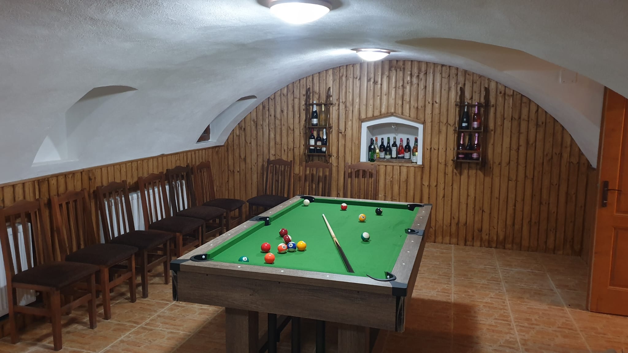 Cramă boltită cu pereți lambrisați din lemn, masă de biliard și rafturi cu sticle de vin