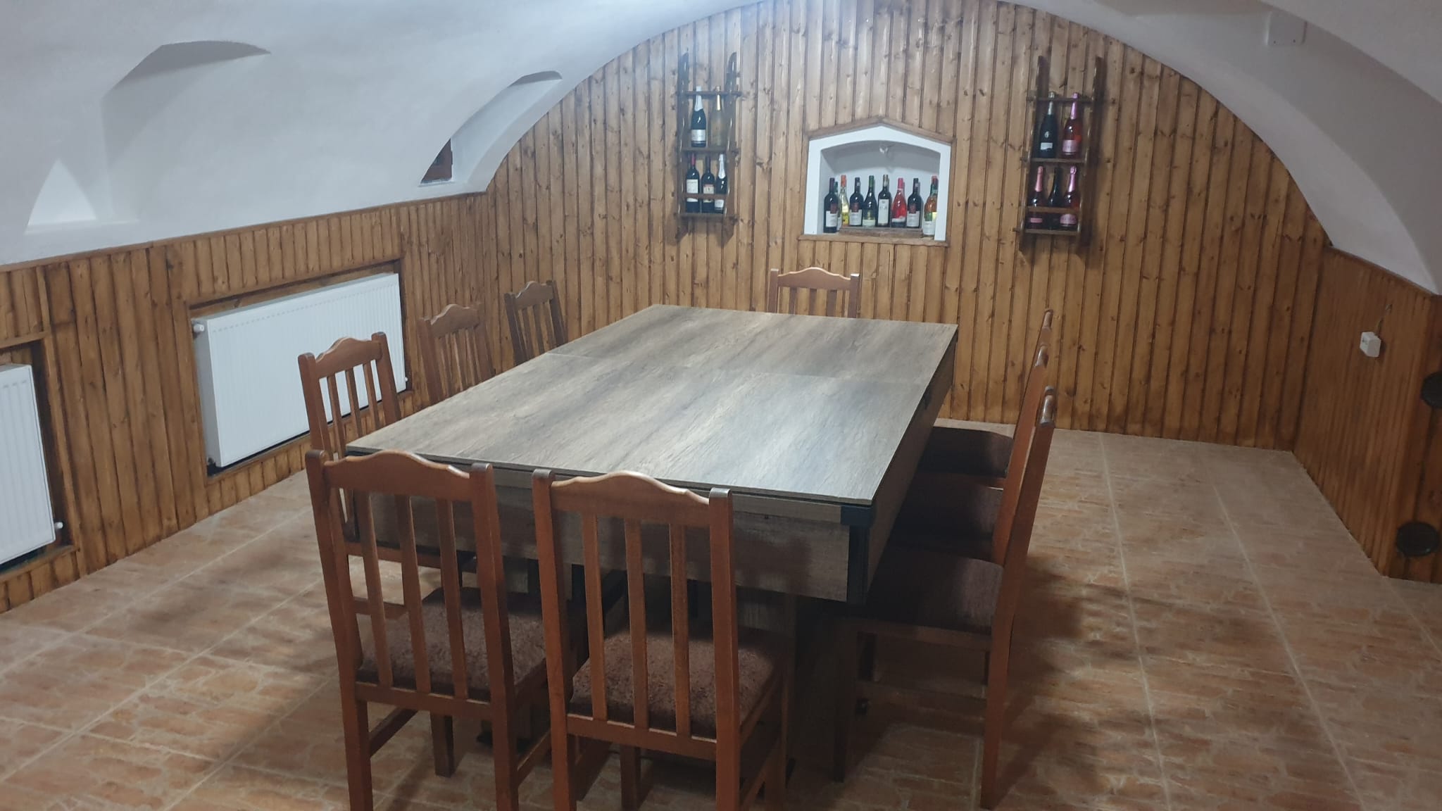 Cramă boltită cu masă pătrată din lemn, scaune și rafturi cu sticle de vin